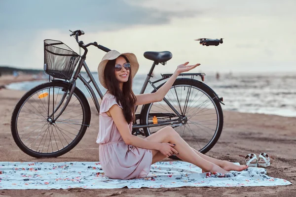 Portret van een jonge brunette meisje in zonnebril en hoed dragen jurk genieten van de vakantie op het strand, spelen met een quadcopter. — Stockfoto