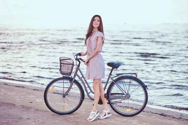 Sonriente chica encantadora con vestido camina con su bicicleta en la playa . — Foto de Stock