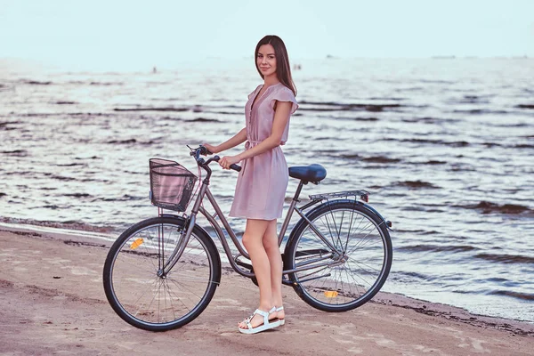 Sonriente chica encantadora con vestido camina con su bicicleta en la playa . — Foto de Stock