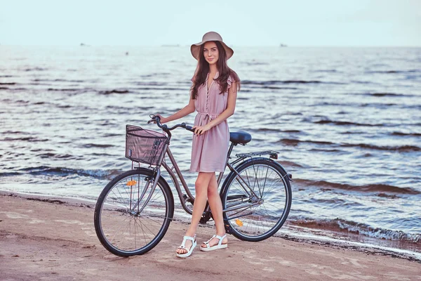 Gelukkig sensuele meisje in hoed dragen jurk loopt met haar fiets op het strand tegen geweldige badplaats achtergrond. — Stockfoto