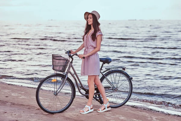 Gelukkig sensuele meisje in hoed dragen jurk loopt met haar fiets op het strand tegen geweldige badplaats achtergrond. — Stockfoto