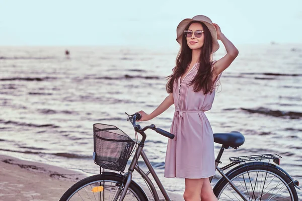 Glad sensuell tjej i solglasögon och hatt bär klänning går med sin cykel på stranden mot en fantastisk kust-bakgrund. — Stockfoto