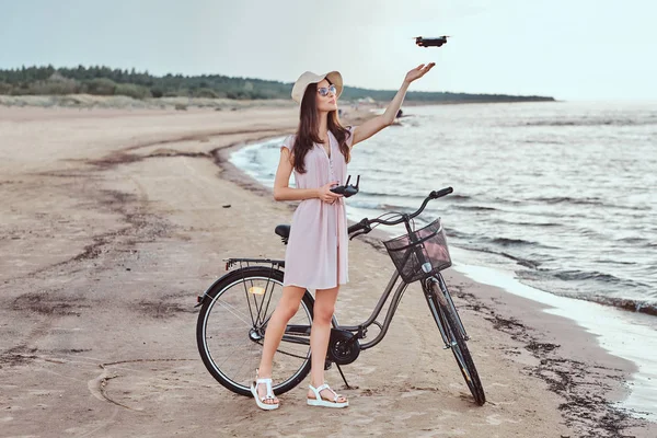 Encantadora chica en gafas de sol y sombrero con vestido disfrutar de las vacaciones en la playa, jugando con el quadcopter . — Foto de Stock