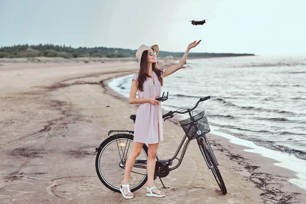 Charming girl in sunglasses and hat wearing dress enjoy the vacation on the beach, playing with the quadcopter. — Stock Photo, Image