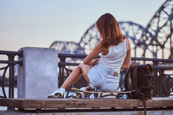 Vista posterior de una niña patinadora vestida con pantalones cortos y camiseta sentada en el terraplén durante el hermoso atardecer . — Foto de Stock