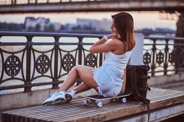 Back view of a skater girl dressed in shorts and t-shirt sitting on the embankment during beautiful sunset. — Stock Photo, Image