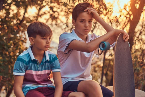 Retrato de dos hermanos pequeños con un monopatín sentado en la barandilla de piedra al aire libre al atardecer . —  Fotos de Stock