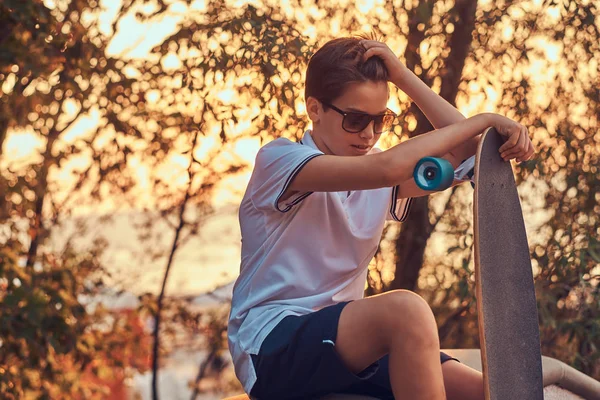 Mladý skejťák v sluneční brýle na sobě tričko a šortky sedí na kamenné zábradlí venku při západu slunce. — Stock fotografie