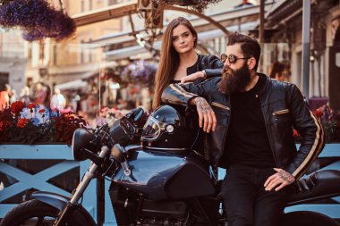 Çekici hipster çift - sakallı bir kafe Teras karşı poz vahşi erkek güneş gözlüğü giymiş bir siyah deri ceket ve onun genç seksi kız yanında, ayakta.
