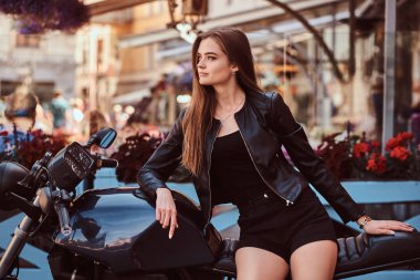Şehvetli genç kız modeli bir siyah elbise ve bir kafe Teras karşı poz özel yapım bir motosiklet üzerinde oturan deri ceket giymiş.
