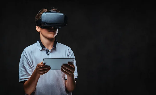 Portret van een jonge student jongen gekleed in een wit t-shirt met behulp van virtual reality bril en tablet pc. — Stockfoto