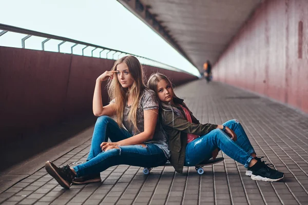 母亲和她的女儿的金发女郎穿着时髦的衣服坐在一起在一个滑板在桥人行道 — 图库照片