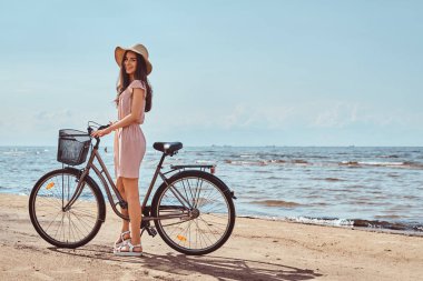 Mutlu çekici kız elbise ve şapka sahilde bisiklet ile güneşli bir günde poz giyinmiş.