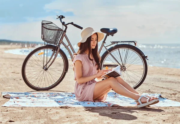 Charming young woman holds a book and looking at camera while sitting on the beach.
