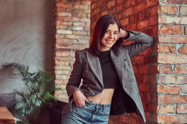 Gülümseyen şehvetli esmer kadın çatı iç ile bir odada bir tuğla duvara yaslanmış, kameraya bakarak trendy kıyafetler giymiş.