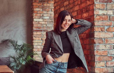 Gülümseyen şehvetli esmer kadın çatı iç ile bir odada bir tuğla duvara yaslanmış, kameraya bakarak trendy kıyafetler giymiş.