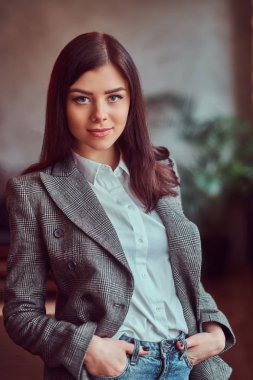 Yakın çekim portre sevimli bir esmer kadının elleri ceplerinde ile poz çatı iç ile bir odada bir masada yaslanmış, kameraya bakarak bir gri şık ceket giymiş.