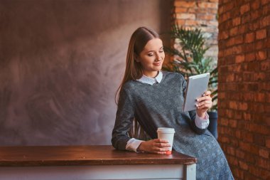 Genç kız zarif bir gri elbise giymiş bir fincan paket servisi olan restoran kahve ve bir oda çatı iç tablo yaslanmış bir tablet kullanarak tutar.
