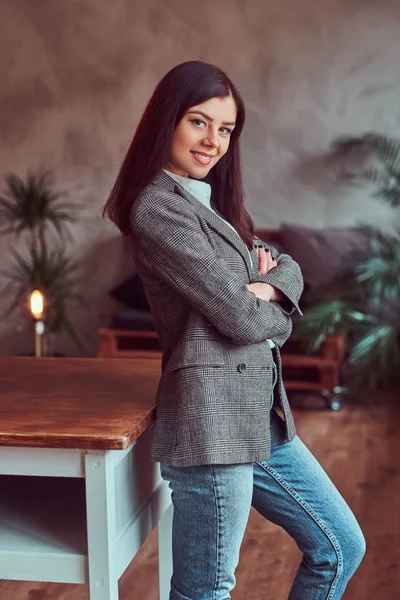 ロフトのインテリアの部屋のテーブルの上に傾いて カメラを見てポーズ グレー エレガントなジャケットに身を包んだ幸せな若いブルネットの女性の肖像画 — ストック写真