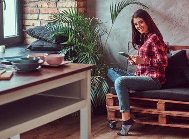Gülümseyen esmer kız öğrenci bir ekose gömlek ve çatı iç ile bir odada bir kanepede oturmuş, kameraya bakarak dijital tablet tutan kot giymiş.