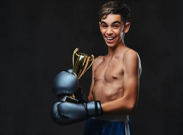 手袋を着用してうれしそうな若い上半身裸ボクサー チャンピオンは優勝カップを保持します 暗い背景に分離 — ストック写真