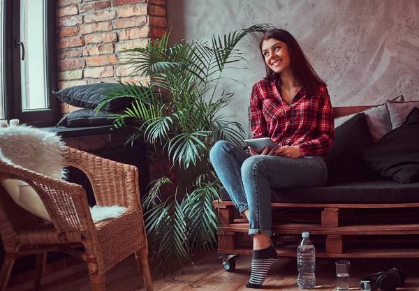 格子縞のシャツとロフトのインテリアと部屋のソファに座って 窓の外見ているデジタル タブレットを保持しているジーンズを着て幸せなブルネットの女の子学生 — ストック写真