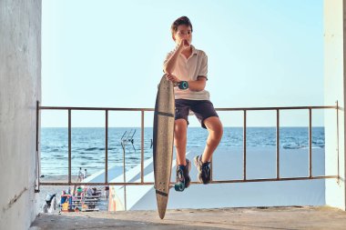 Dalgın genç kaykaycı çocuk portresi t-shirt ve şort Otokorkuluk üzerinde oturan giymiş ve bir kaykay sahil arka planı tutar.