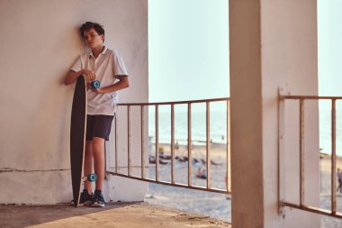 Bir genç kaykaycı çocuk portresi t-shirt ve şort bir duvara yaslanmış giymiş ve bir kaykay bir sahil arka planı tutar.