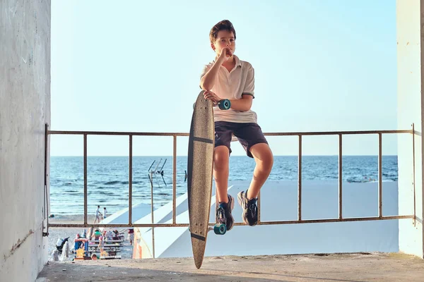 Dalgın genç kaykaycı çocuk portresi t-shirt ve şort Otokorkuluk üzerinde oturan giymiş ve bir kaykay sahil arka planı tutar.
