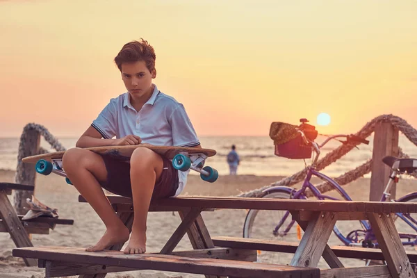 シャツと明るい夕日を背景に 海岸のベンチに座ってパンツに身を包んだ若いスケーターの少年 — ストック写真