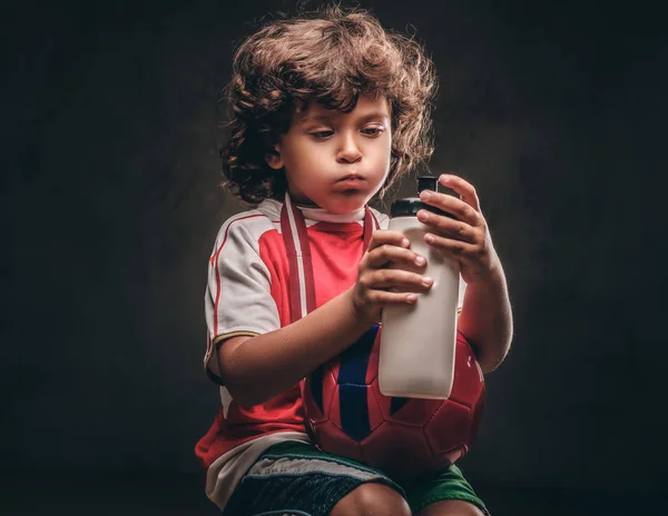 Little champion boy in sportswear holds a ball and drinking water from a bottle. Isolated on the dark textured background.