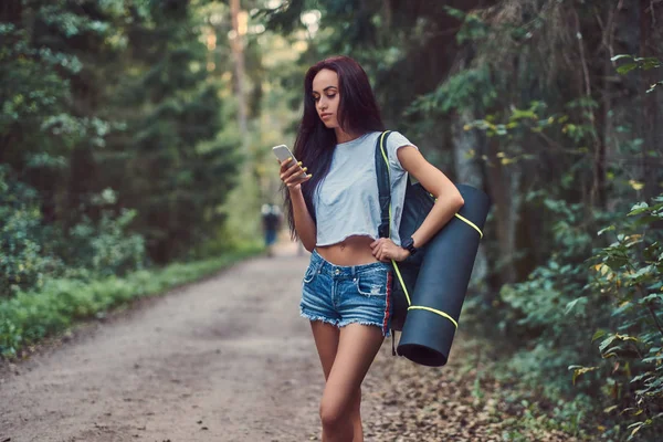 迷人的时髦女孩穿着衬衫和短裤与旅游垫和背包使用智能手机 而站在夏季森林 — 图库照片