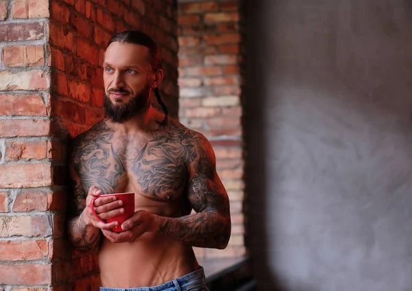 Elegante hombre barbudo sin camisa con cuerpo musculoso y tatuado sostiene la taza de café mientras se apoya en una pared y mira por la ventana . —  Fotos de Stock