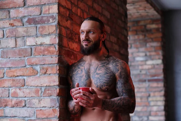 Eleganta bar överkropp skäggiga män med muskulös och tatuerade kropp håller kopp kaffe medan lutande på en vägg och ser ut genom fönstret. — Stockfoto