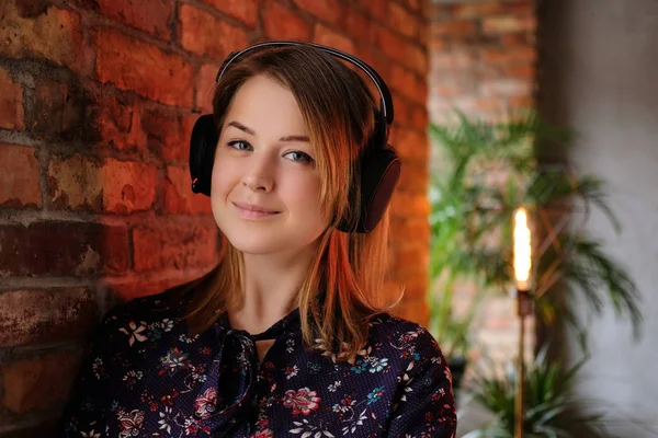 Happy blonde woman in dress listening to music in headphones while leaning on a brick wall in room with loft interior. — Stock Photo, Image