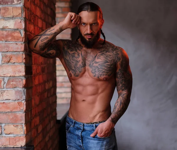 Pensive shirtless bearded male with the muscular and tattooed body leaning on a wall and looks at the camera. — Stock Photo, Image