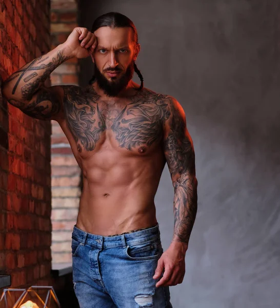 Pensivo sem camisa barbudo macho com o corpo musculoso e tatuado encostado em uma parede e olha para a câmera . — Fotografia de Stock