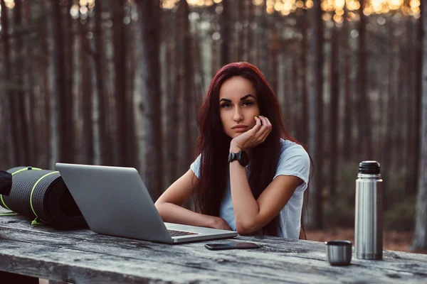 Chica hipster pensativo en camisa blanca mirando a la cámara mientras está sentado en un banco de madera con un ordenador portátil abierto en el hermoso bosque de otoño . — Foto de Stock