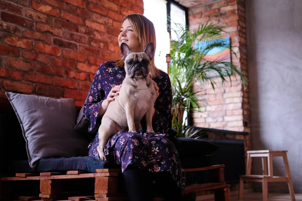 Mujer rubia feliz en vestido sentado con su lindo pug en un sofá hecho a mano en la habitación con interior loft . —  Fotos de Stock