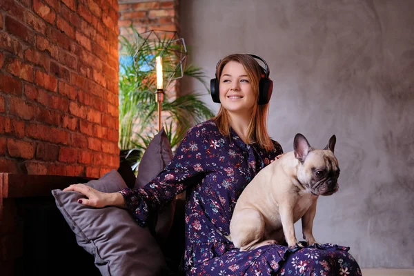 Mulher loira feliz no vestido sentado com seu pug bonito em um sofá artesanal e ouvir música em uma sala com interior loft . — Fotografia de Stock