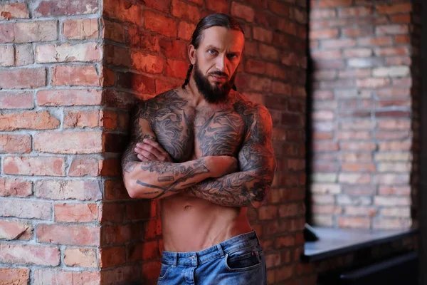 Elegante hombre barbudo con cuerpo musculoso y tatuado apoyado en una pared con los brazos cruzados y mirando a la cámara . —  Fotos de Stock