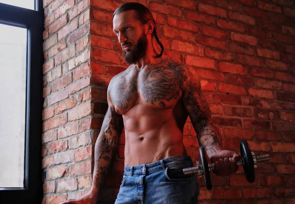 Bărbat tatuat cu barbă brutală care face exerciții fizice cu o gantere într-o cameră cu interior mansardă . — Fotografie, imagine de stoc