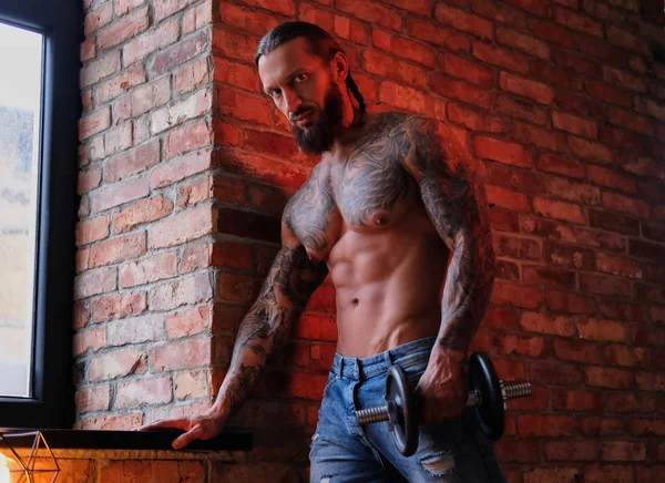 Brutal bebaarde tattooed man doen oefening met een halter in een kamer met loft interieur. — Stockfoto