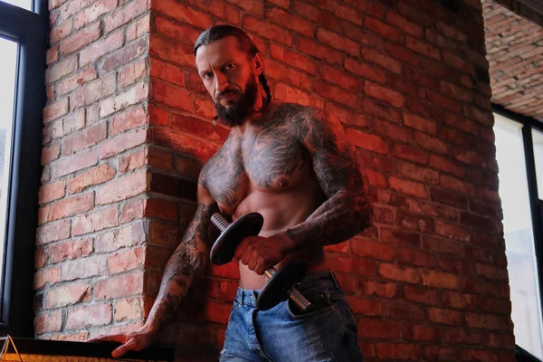 Жорстокий бородатий татуйований чоловік робить вправи з гантелі в кімнаті з інтер'єром лофт . — стокове фото