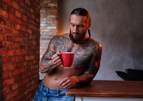 Varón barbudo con cuerpo tatuado sostiene una taza de café mientras se apoya en una mesa en una habitación con interior loft . — Foto de Stock