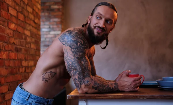 Maschio barbuto sorridente con corpo tatuato tiene la tazza di caffè mentre si appoggia su un tavolo in una stanza con interno soppalco . — Foto Stock