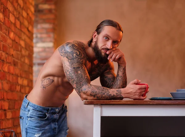 Töprengő szakállas férfi, a tetovált test tart csésze kávét, miközben támaszkodva egy asztal-ban egy szoba, loft belső. — Stock Fotó