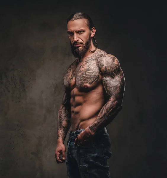 Retrato de un hombre sin camisa con barba tatuado posando con la mano en el bolsillo. Aislado sobre fondo de textura oscura . —  Fotos de Stock