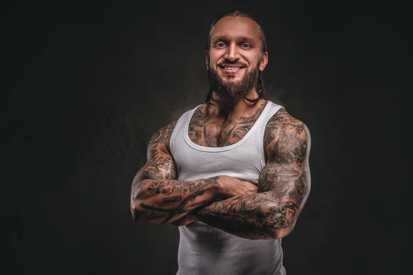 Glimlachend knappe bebaarde tattooed man in wit overhemd poseren met gekruiste armen. Geïsoleerd op een donkere gestructureerde achtergrond. — Stockfoto