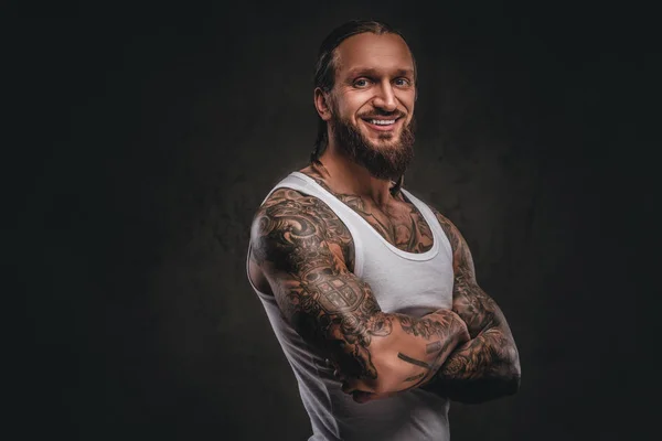 Smiling handsome bearded tattooed male in white shirt posing with crossed arms. Isolated on a dark textured background. — Stock Photo, Image